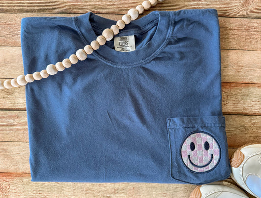 Blue Comfort Colors pocket t-shirt with a smiley face pocket patch on a wooden surface
