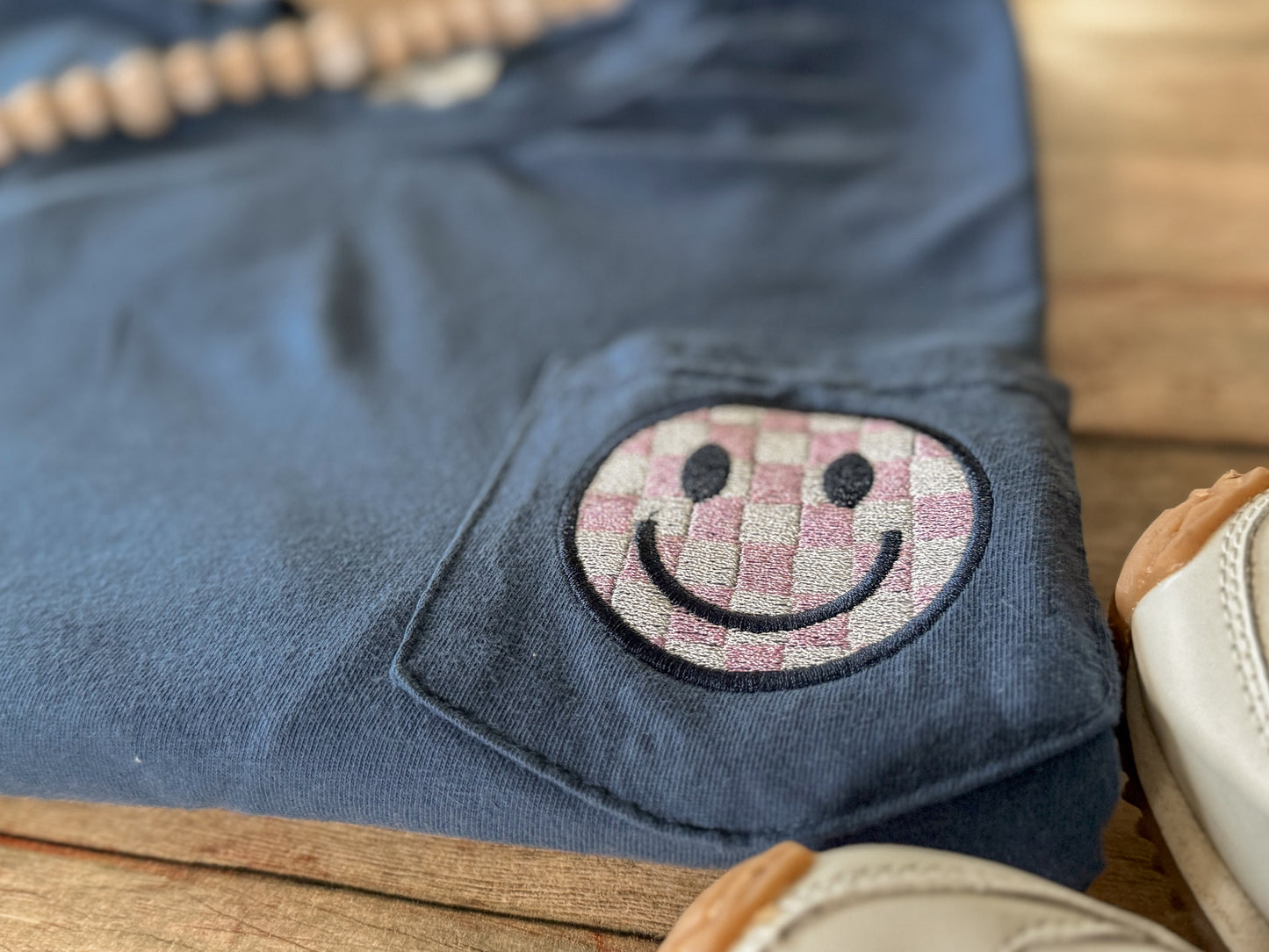 Blue Comfort Colors pocket tee with a smiley face patch on a wooden surface