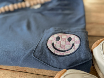 Blue Comfort Colors pocket tee with a smiley face patch on a wooden surface